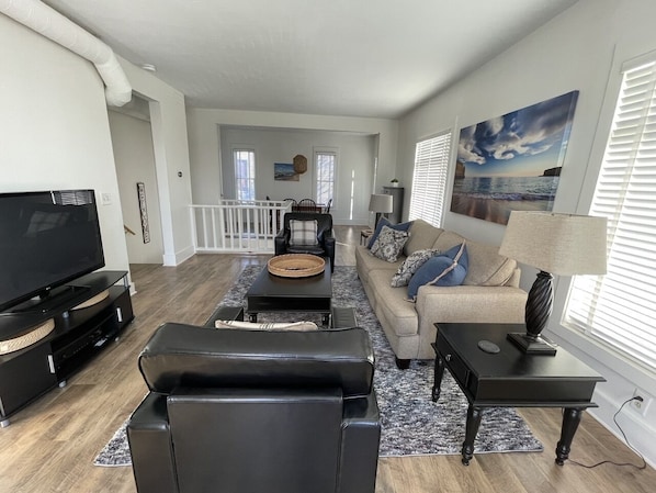 A modern living room with a TV, black leather sofa, and a coffee table. There is a painting on the wall and a window with blinds.