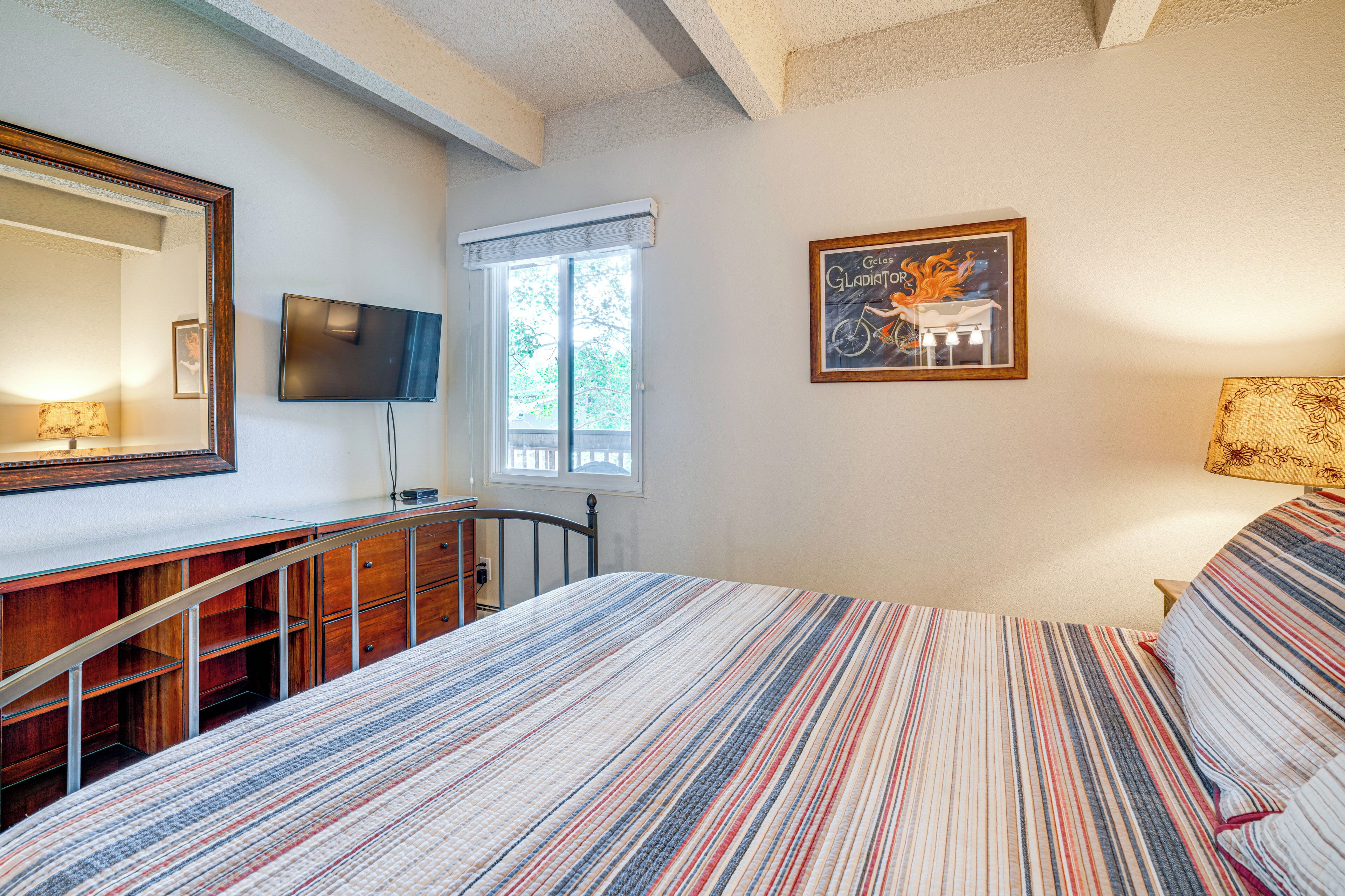A bedroom with a bed, a desk, a chair, a TV, a window, and a framed picture on the wall.