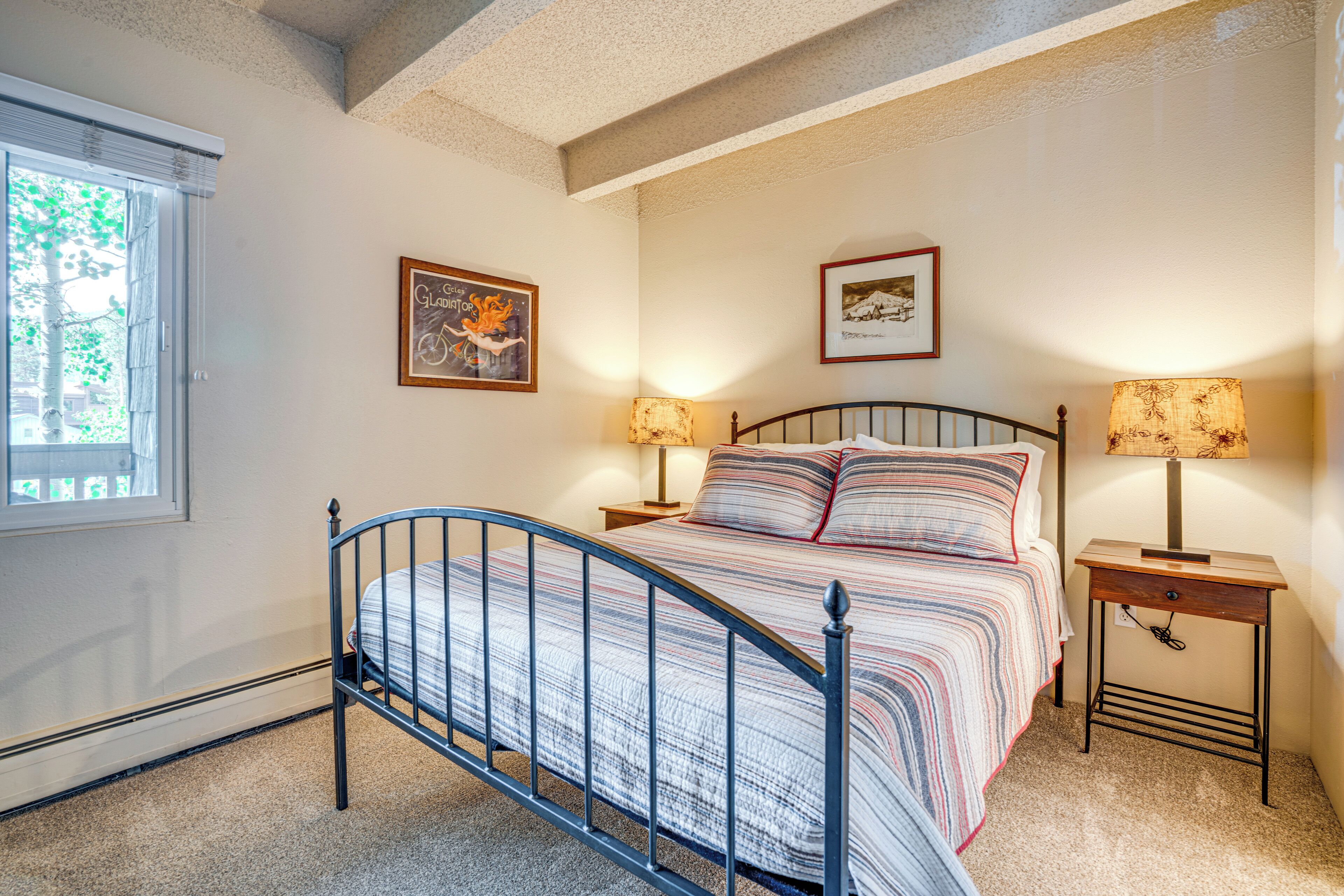 Un dormitorio con estructura de cama metálica, dos mesitas de noche con lámparas, una ventana con persianas y dos cuadros enmarcados en la pared.
