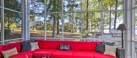 A covered outdoor seating area with a red sofa, a woven coffee table, and a view of trees and a house in the background.
