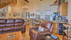 A spacious living room with leather sofas, a wooden coffee table, and a chandelier. A kitchen area with a refrigerator and microwave is visible in the background. - Lakefront Paradise w/ Fire Pit - Dogs Welcome! (Cross Hill)