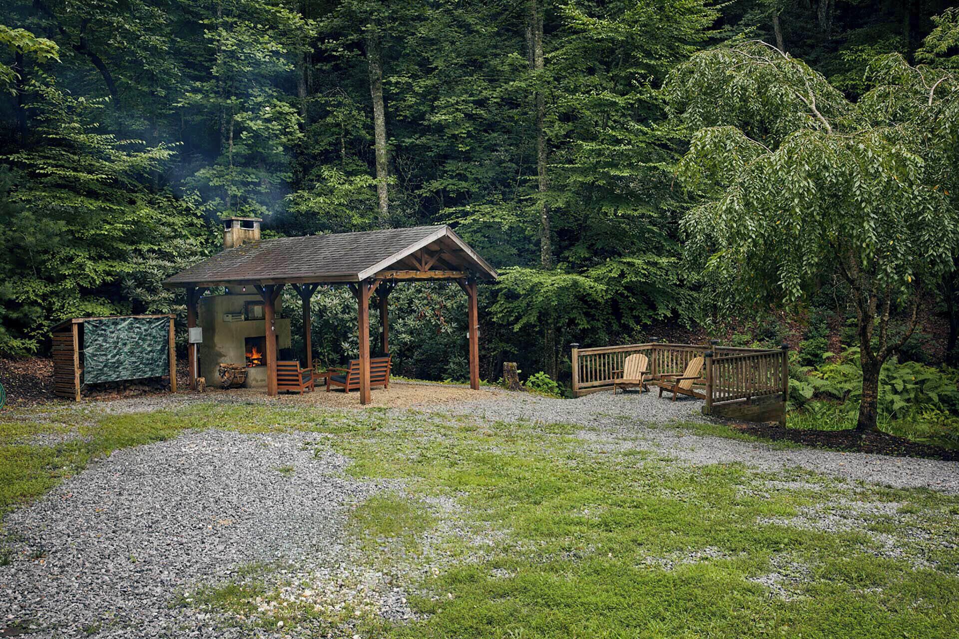 A rustic cabin with a wooden deck, a fire pit, and a covered seating area surrounded by dense forest.
