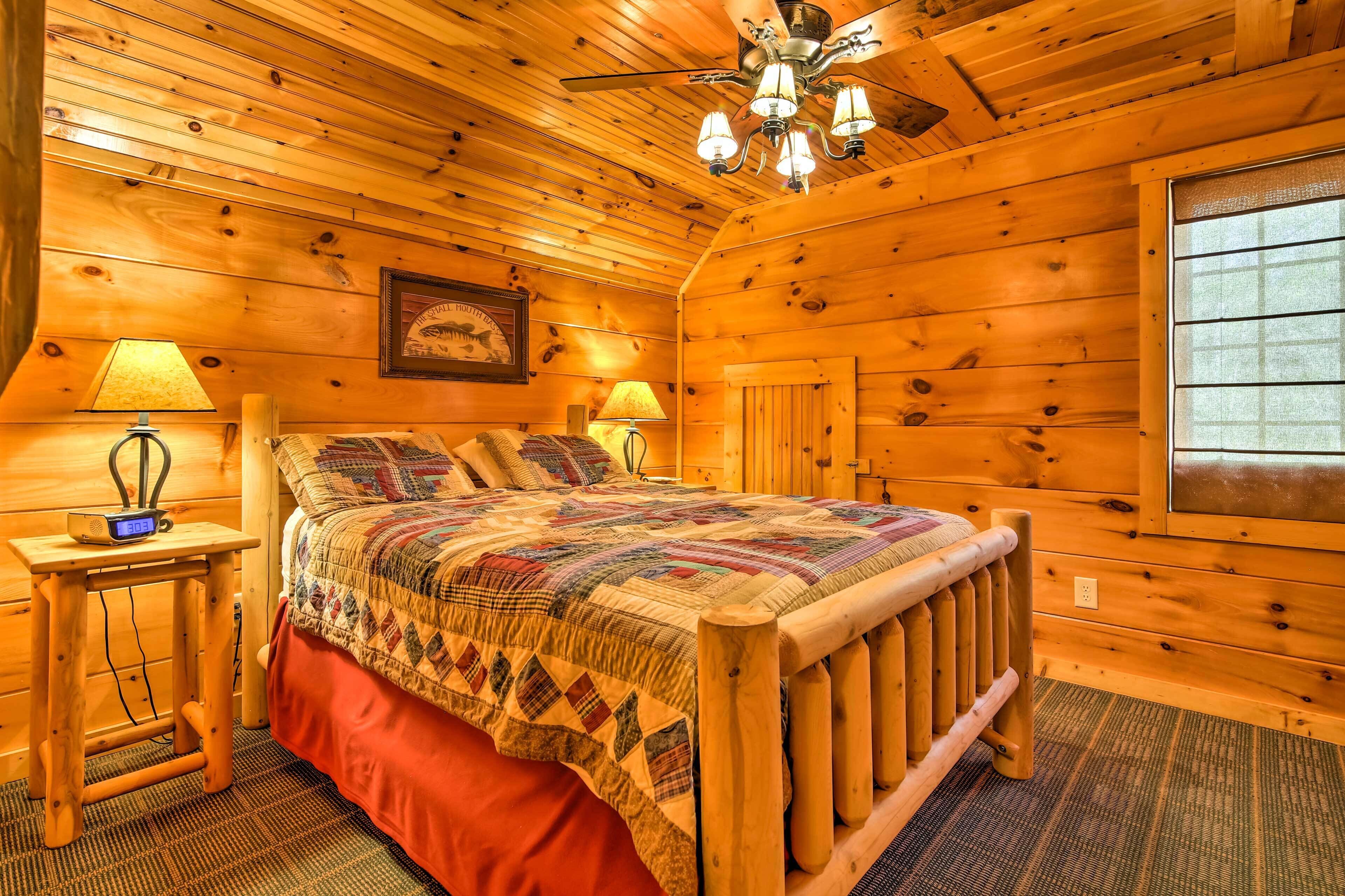 A cozy wooden cabin bedroom with a bed, two bedside tables, a nightstand, a wall-mounted lamp, and a ceiling fan.