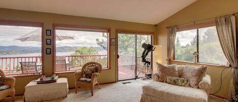 A spacious room with a large window offering a view of a balcony and mountains. There is a comfortable sofa and a chair with a table in the center.
