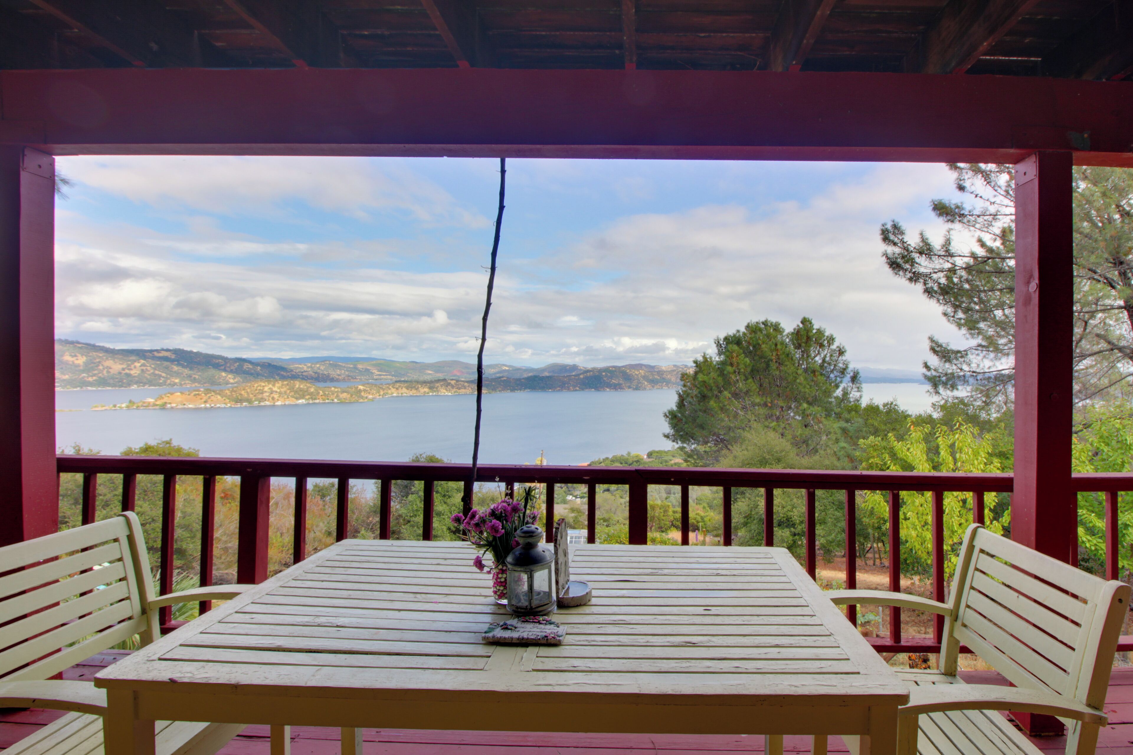 A wooden table with two chairs on a balcony with a red railing, overlooking a lake and hills.