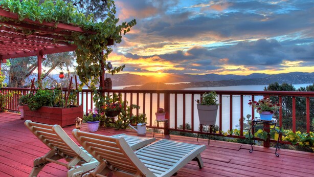 Une terrasse en bois avec deux chaises longues, des plantes en pot et une vue sur un coucher de soleil se reflétant sur une étendue d'eau.