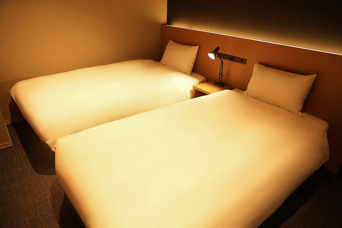 A hotel room with two single beds, a nightstand, and a wall-mounted lamp.