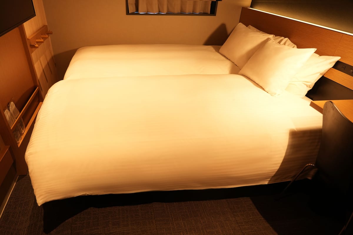 A neatly made bed with white linens and pillows, a wooden headboard, and a bedside table with a lamp.