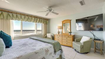 A bedroom with a bed, a dresser, a chair, a television, and a view of the ocean.