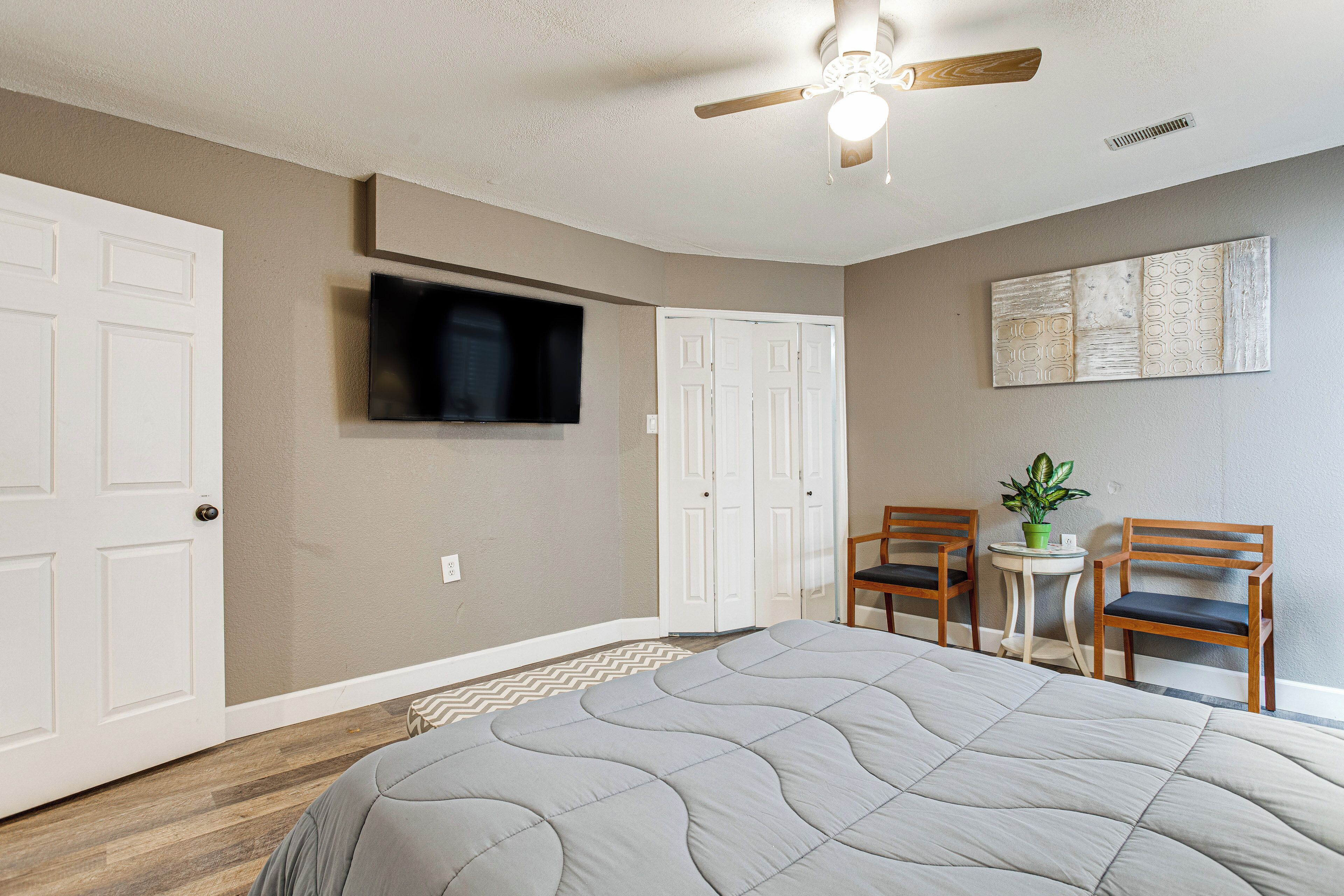 A bedroom with a bed, a ceiling fan, a flat-screen TV, two chairs, a small table with a plant, and a door.