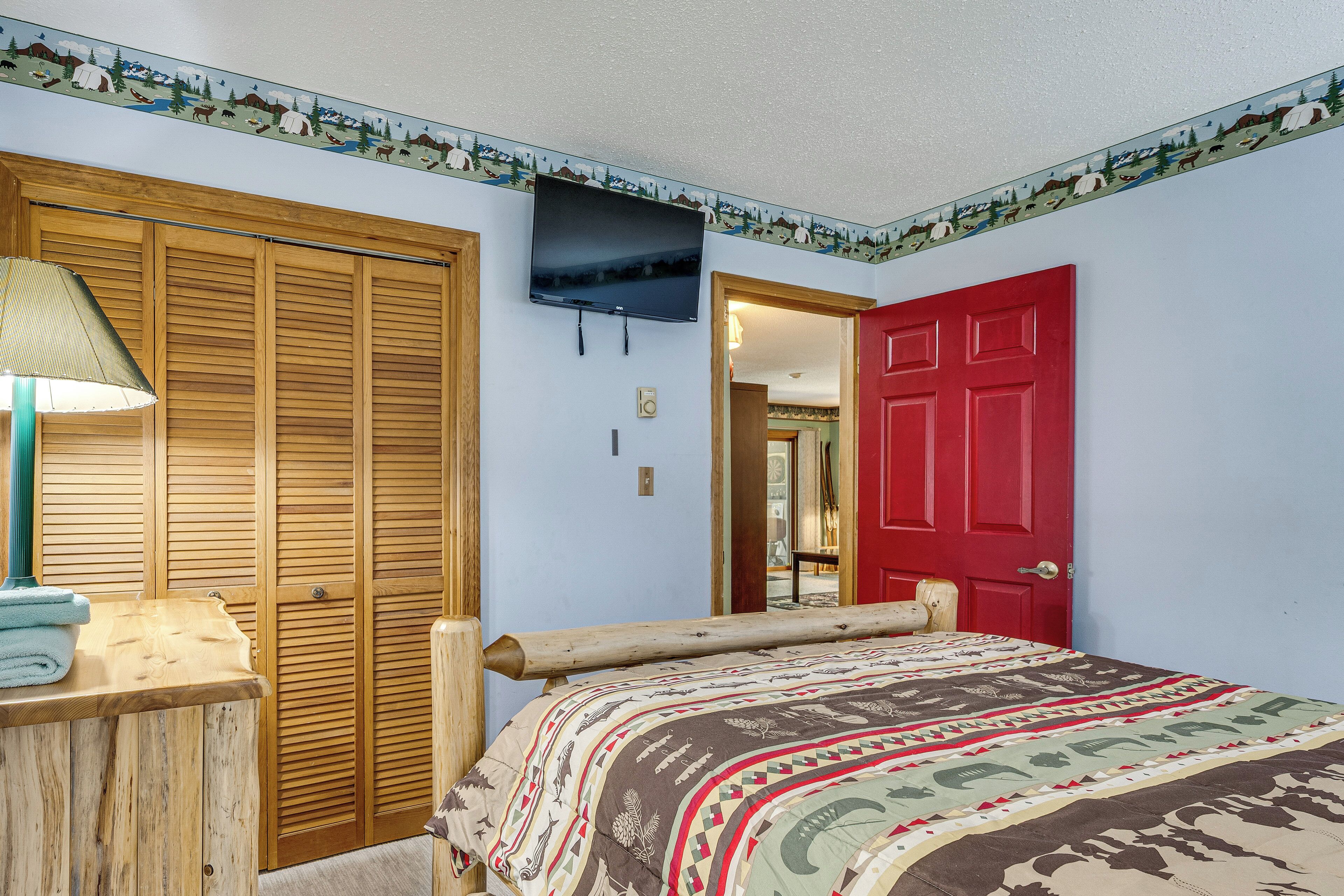 A bedroom with a bed, a TV, wooden closet, a red door, and a lamp.