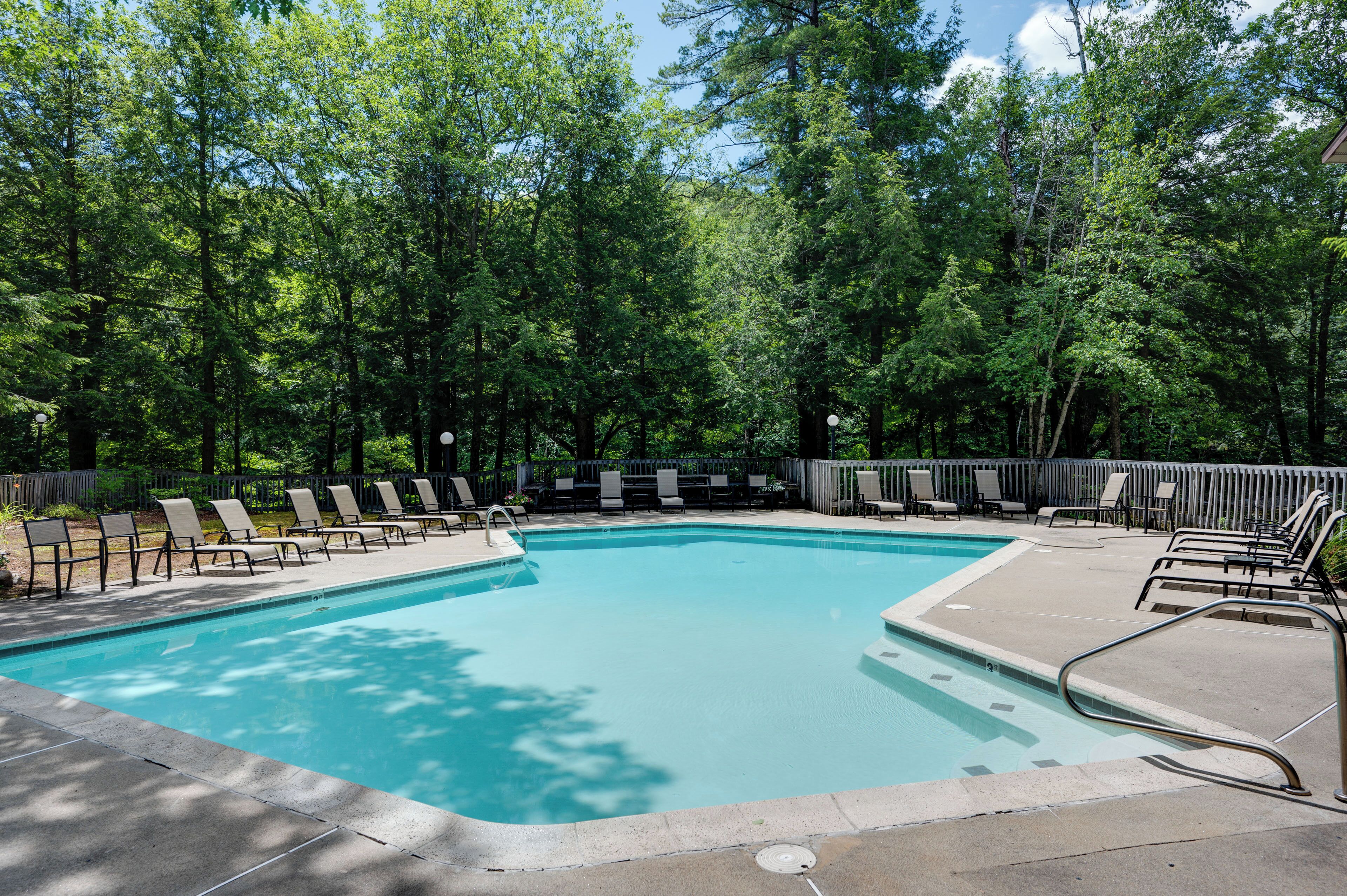 A rectangular pool with a clear blue water surface, surrounded by a concrete deck with lounge chairs and a wooden fence.