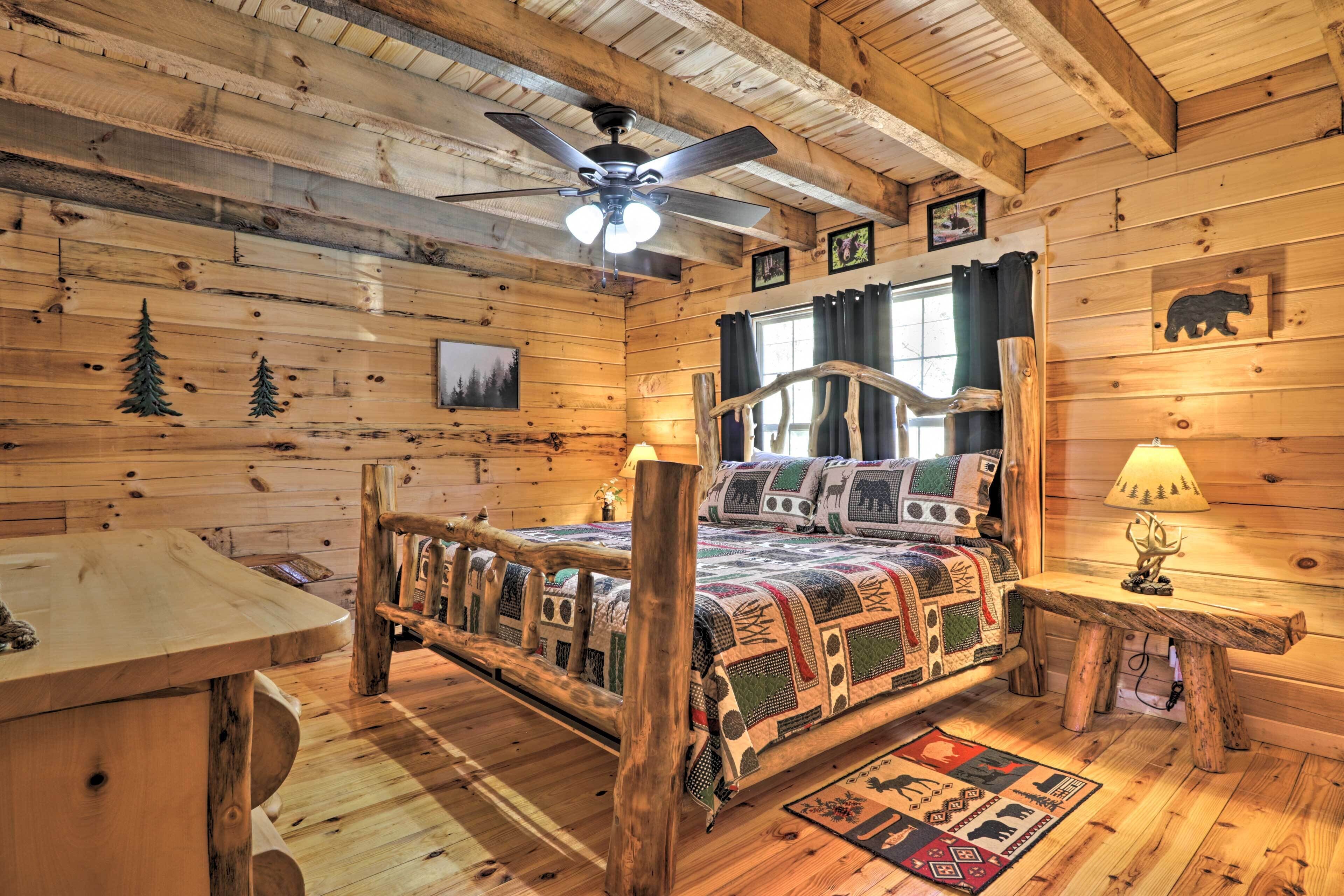Ein Schlafzimmer in einer Blockhütte mit einem hölzernen Bett, Deckenventilator, einem Fenster mit Vorhängen und einem Nachttisch aus Holz mit Lampe.