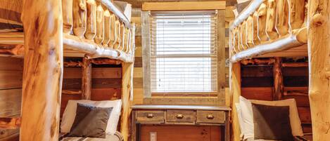 A cabin with two wooden bunk beds, a window with blinds, and a wooden dresser with drawers.