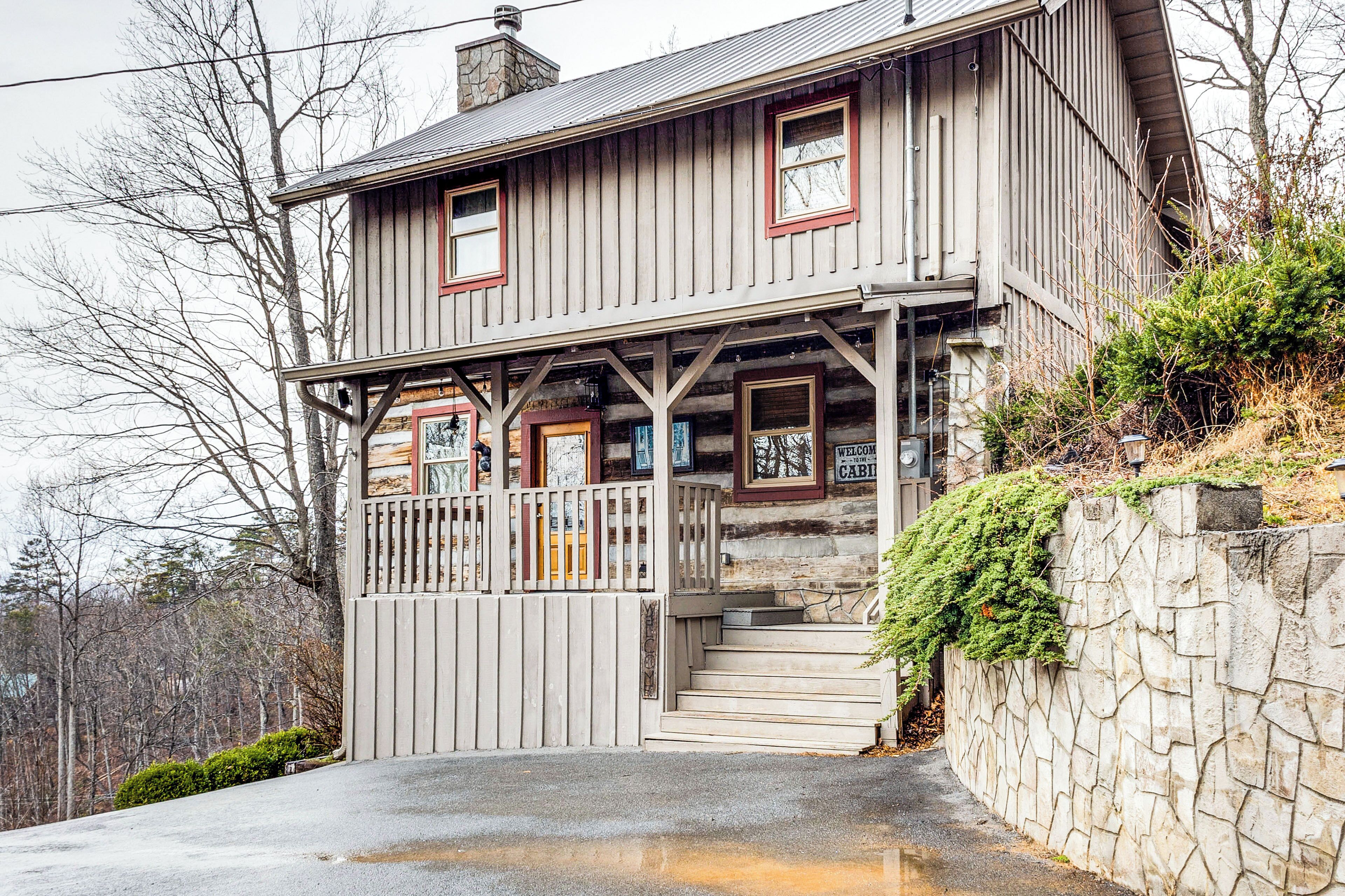 Une cabane en rondins à deux étages, dotée d’une véranda, d’un mur en pierre et d’une pancarte indiquant « Bienvenue à la cabane ».