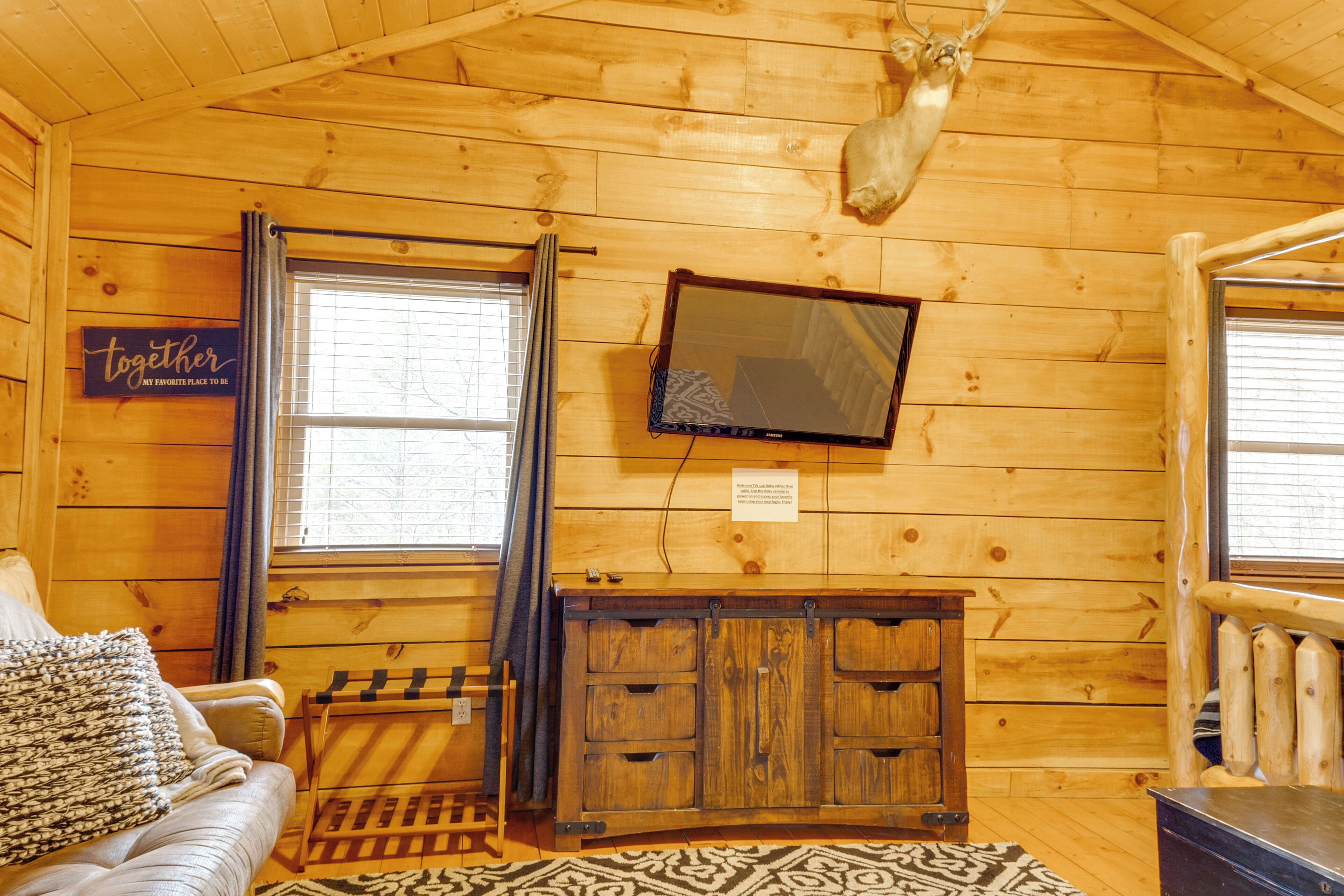 Interior acogedor de una cabaña con piso de madera, un sofá con una manta, un televisor montado y un mueble con cajones.