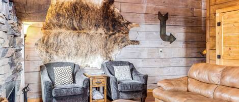 Un salon chaleureux avec une grande moquette en fourrure, deux fauteuils gris, un canapé en cuir marron et une table basse en bois.