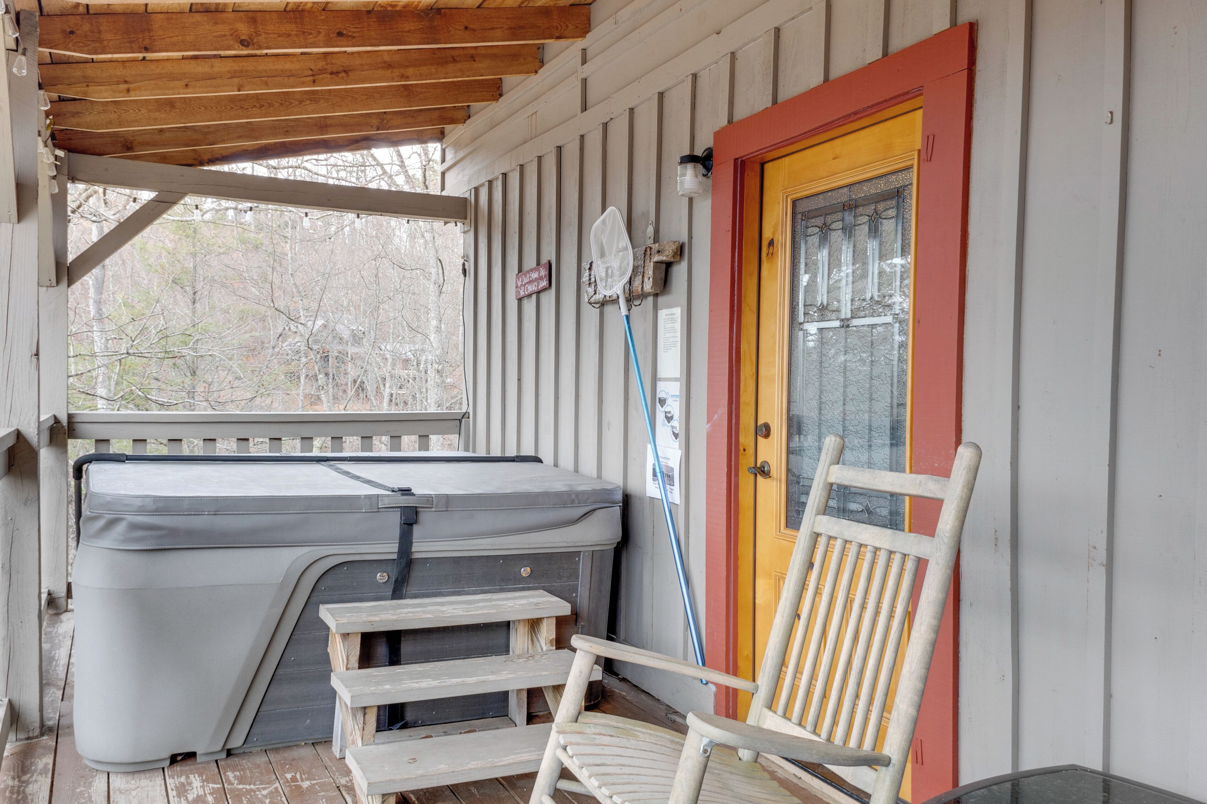 A covered porch with a hot tub, a wooden chair, and a door with a red frame.