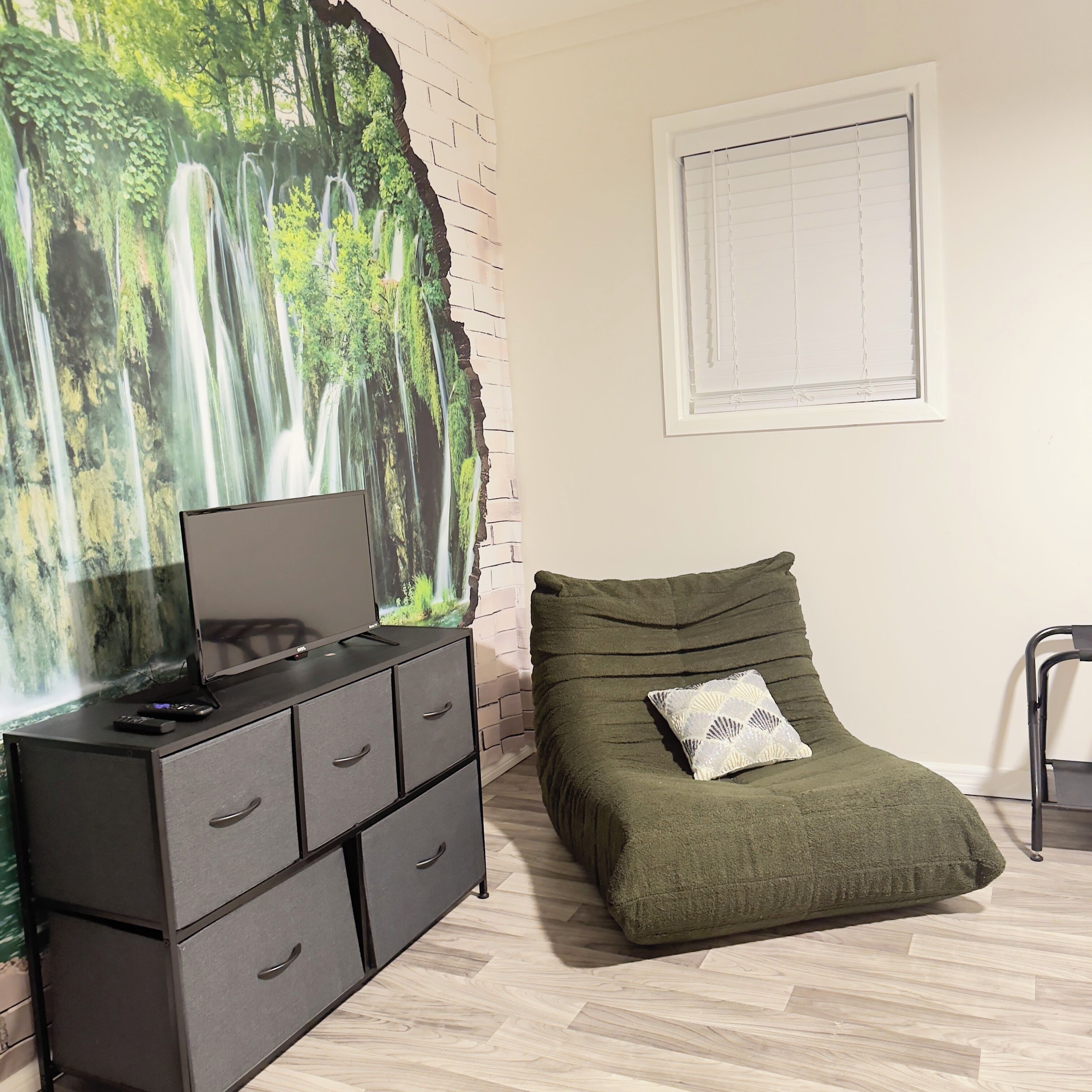 Um quarto com uma cama, uma cômoda com gavetas, uma televisão sobre a cômoda, uma janela com persianas e uma parede com um mural temático da natureza.