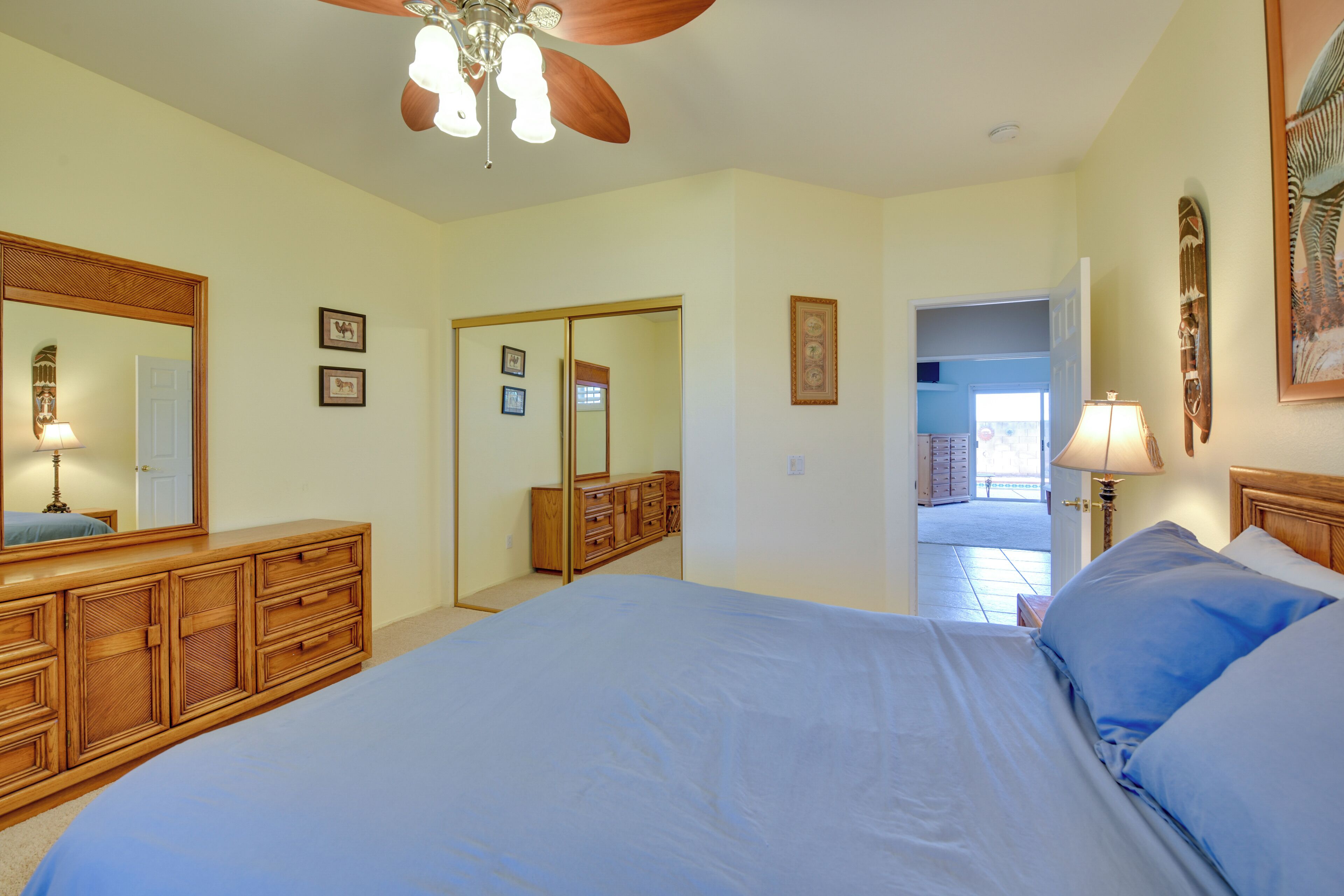 Une chambre avec un grand lit, une commode en bois, un ventilateur de plafond et une porte donnant sur un balcon.