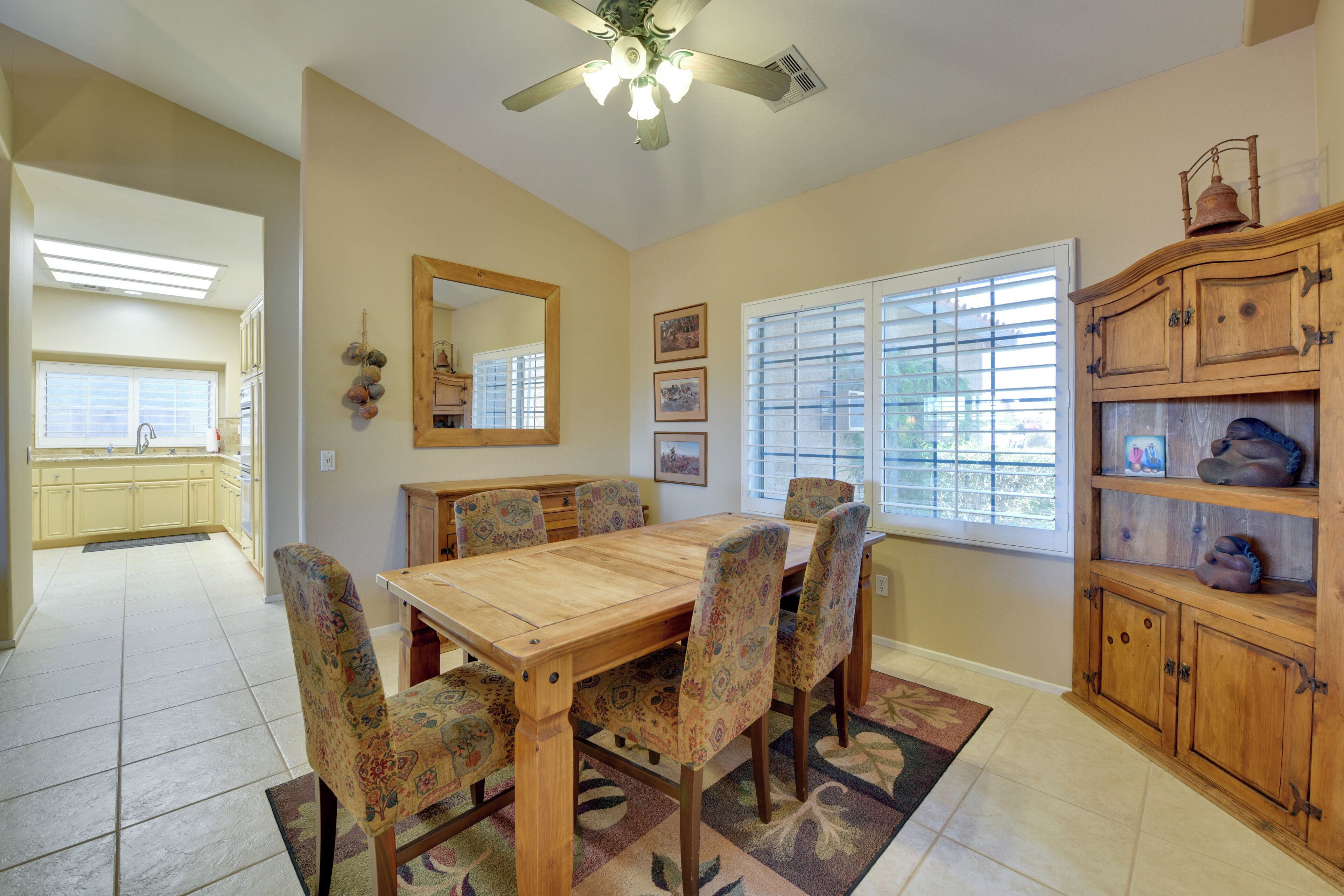 A dining room with a wooden table and patterned chairs, a ceiling fan, and a view to the kitchen.
