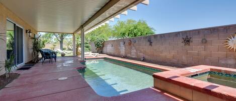 A covered outdoor swimming pool area with a patio, lounge chairs, and a brick wall with decorative elements.