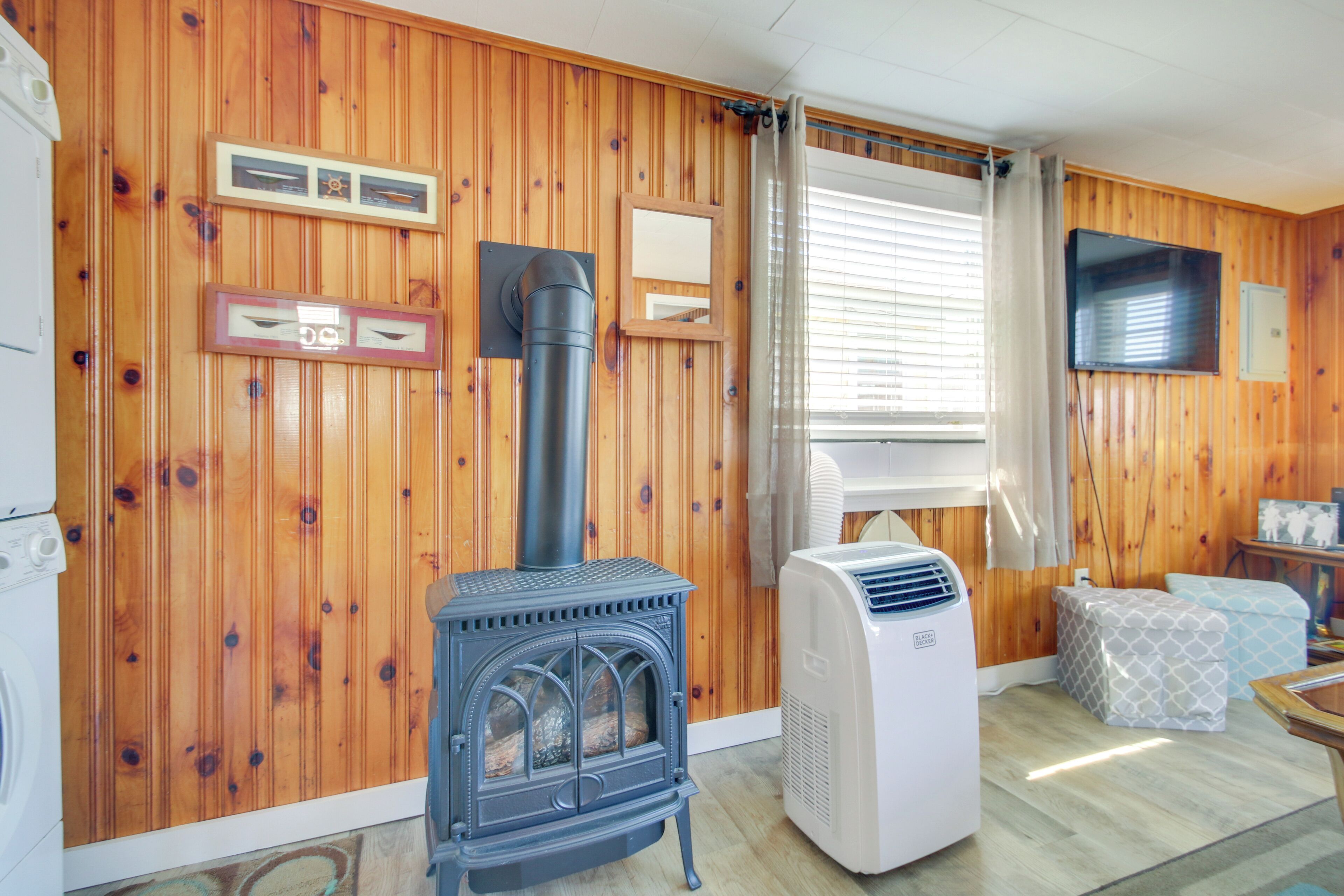 A room with wooden walls, a wood-burning stove, a large air conditioner, a television, and a window with curtains.