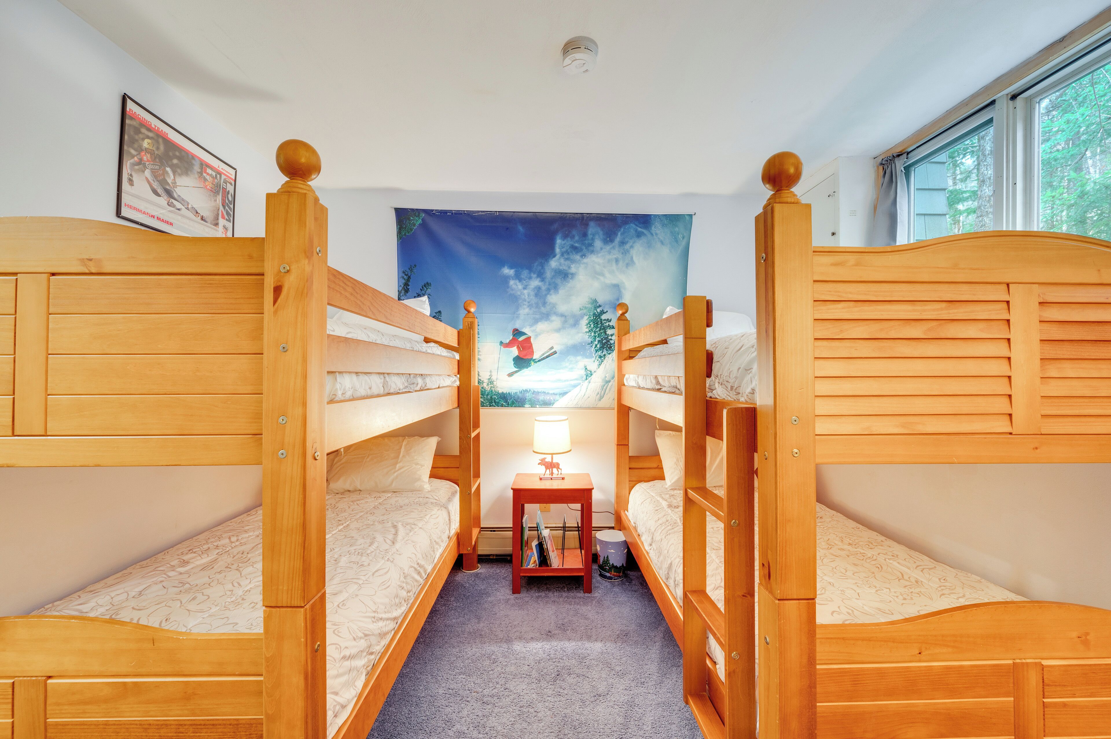 Una habitación con dos camas literas de madera, una mesita con lámpara y un tapiz que muestra a un esquiador.