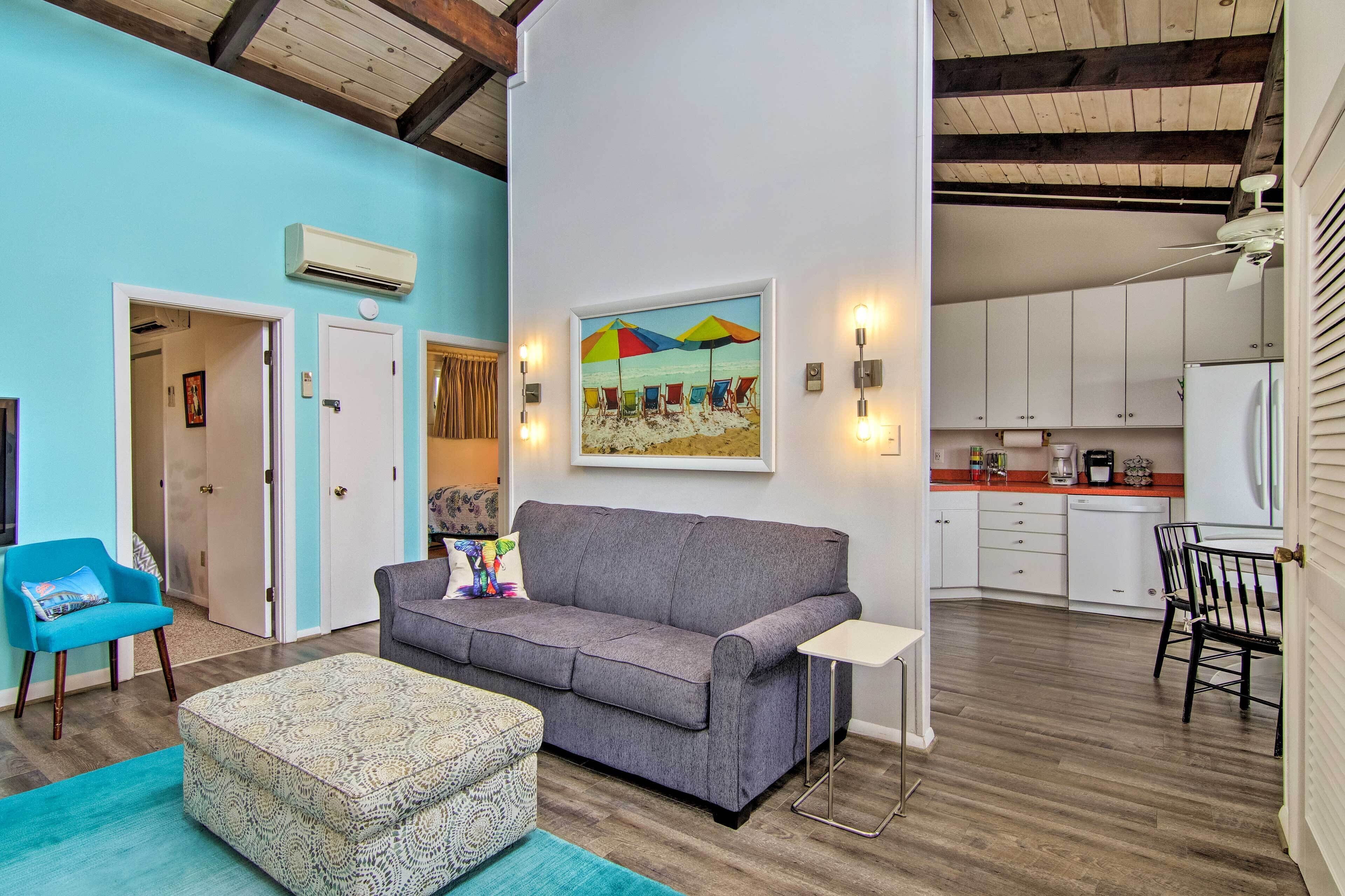 A living room with a grey sofa, a patterned ottoman, a blue armchair, and a painting of beach umbrellas.