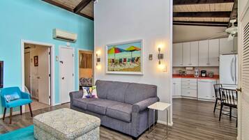 A living room with a grey sofa, a patterned ottoman, a blue armchair, and a painting of beach umbrellas.