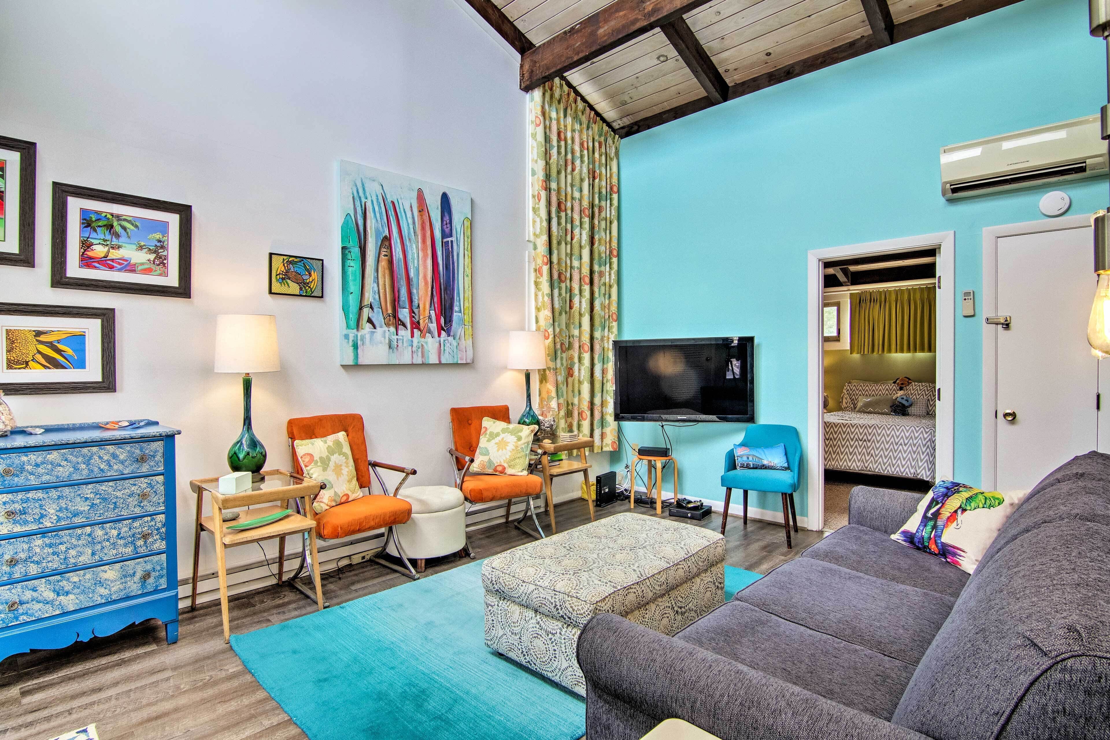 A living room with a sofa, chairs, a coffee table, a TV, and a painting of surfboards.