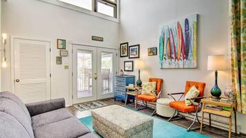 A spacious living room with a gray sofa, a patterned ottoman, and two rocking chairs. A large painting of surfboards hangs on the wall, and there are several framed pictures and a welcome sign above the door.