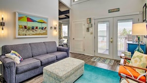 A living room with a gray sofa, a patterned ottoman, and a colorful beach painting. There are two doors leading to a balcony, and a sign that reads "Welcome to the Beach." - Walk to Dewey Beach: Bright Condo w/ Balcony! (Dewey Beach)