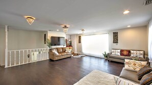 A spacious living room with dark hardwood flooring, a white railing, two sofas with patterned cushions, and a chandelier.