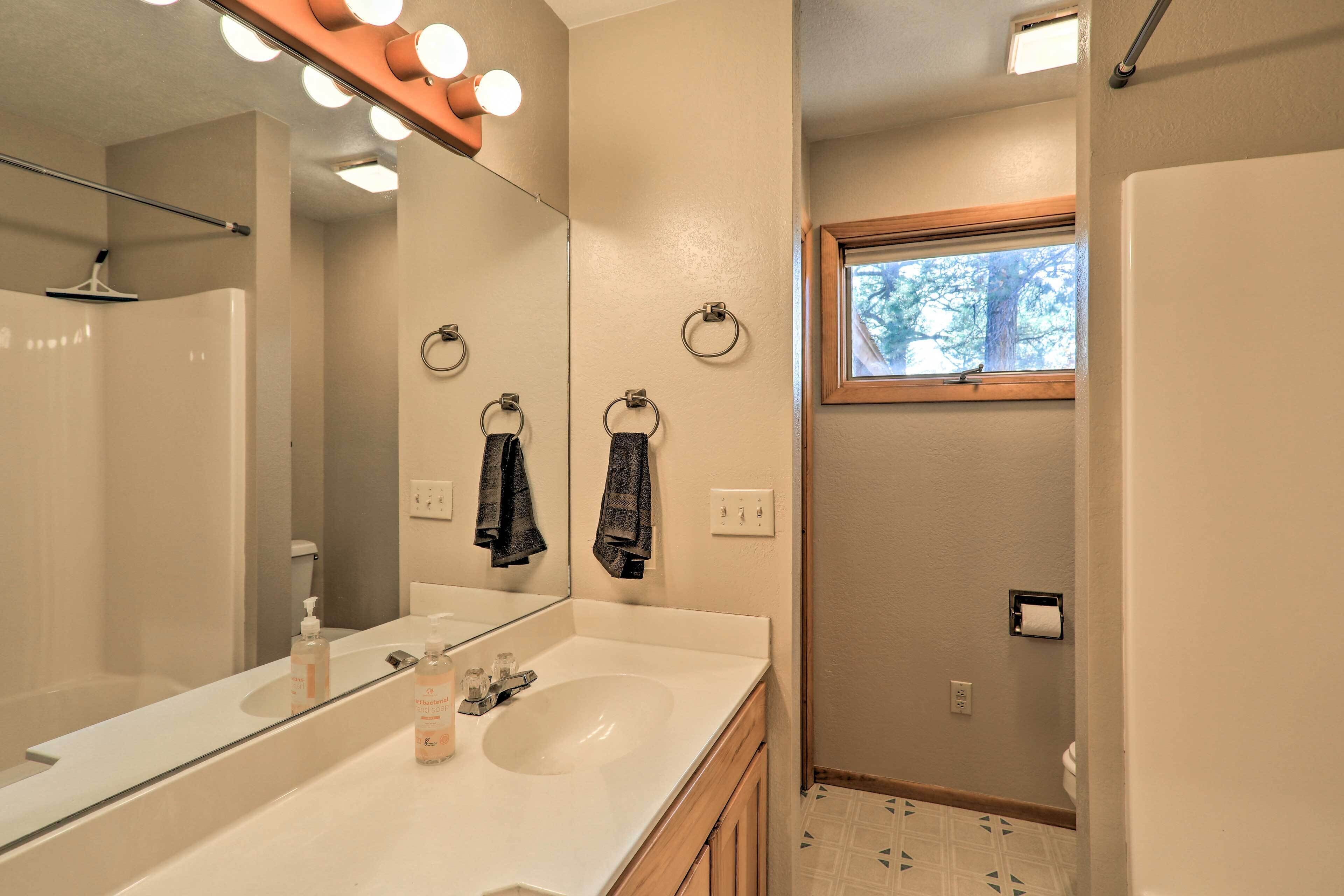 A bathroom with a large mirror, a sink with soap dispensers, a towel ring, and a window with a view of trees.