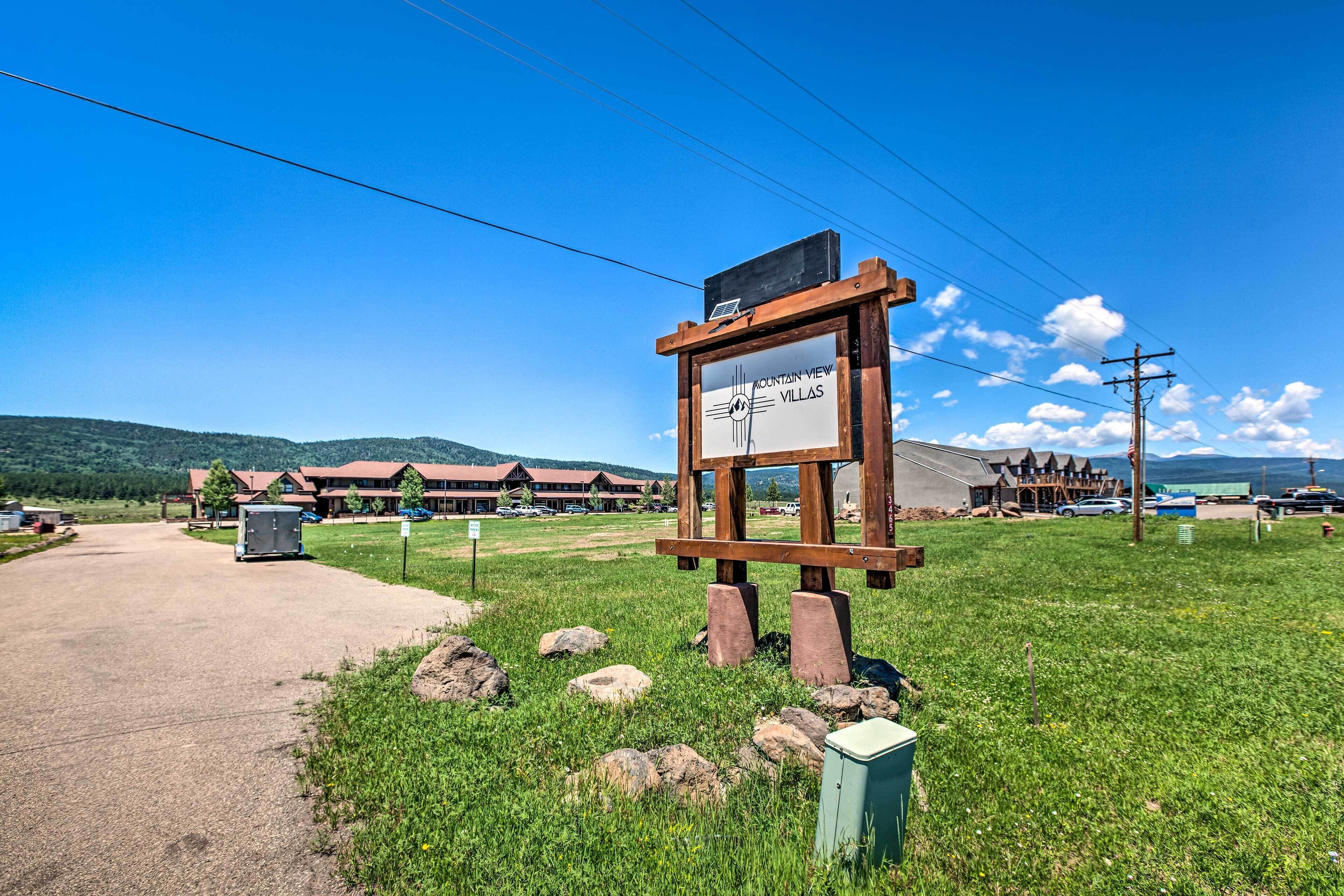 A sign for Mountain View Villas with a clear view of the surrounding area, including a building with a red roof and a dirt road.