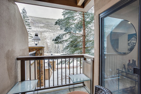 A balcony with a glass table, a wicker chair, and a view of a snowy landscape with trees and a mountain.