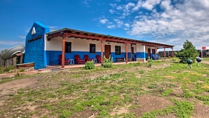 A two-story building with a blue and white facade, a covered porch with red chairs, and a satellite dish on the left. - Chic Studio w/ Grill - 25 Mins to Taos Ski Valley! (El Prado)