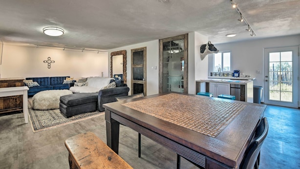 A spacious living area with a large wooden table, a blue sofa, and a kitchen area with a glass door. - Chic Studio w/ Grill - 25 Mins to Taos Ski Valley! (El Prado)