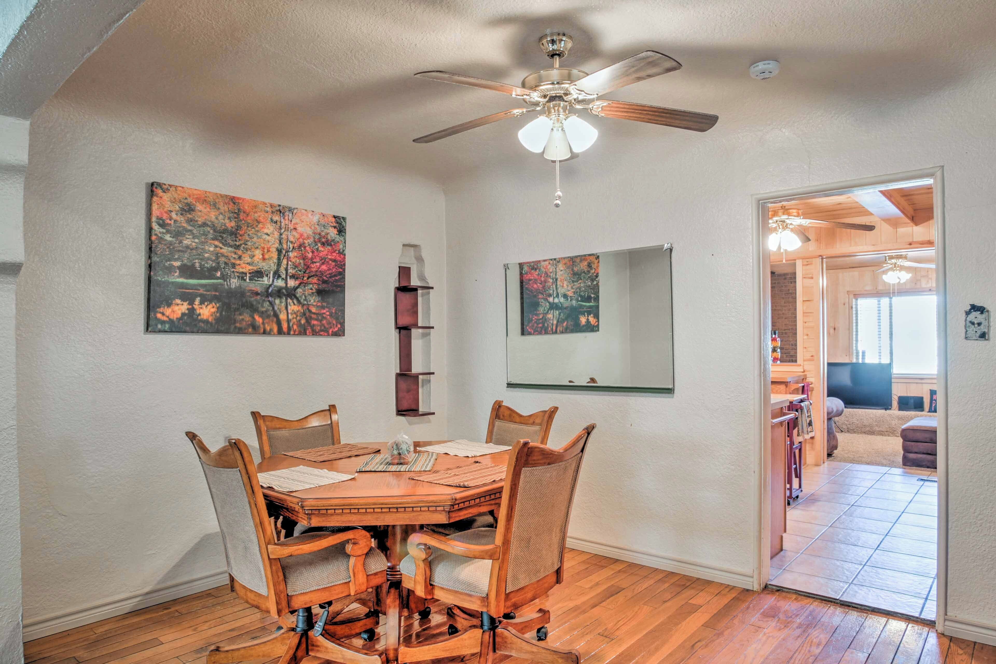 A dining area with a wooden table and chairs, a ceiling fan, and two paintings on the walls.