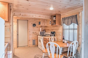 A compact kitchen with wooden walls, a microwave, stove, and white chairs around a table.