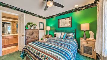A bedroom with a green wall, a bed with patterned bedding, a wooden nightstand, a ceiling fan, and a bathroom visible through an open door.