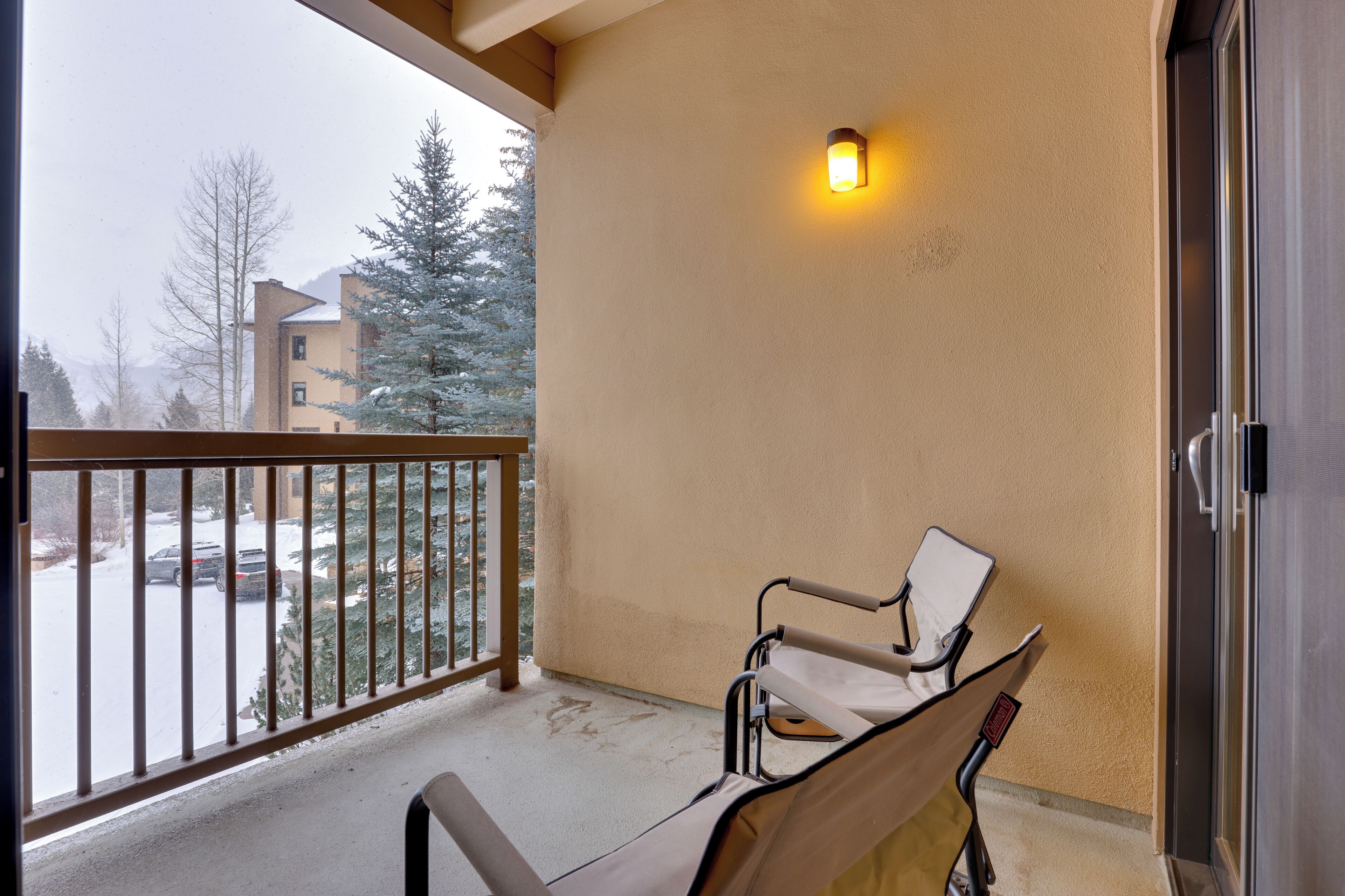 Un balcón con una silla, una luz empotrada en la pared y vistas a un paisaje nevado con edificios.