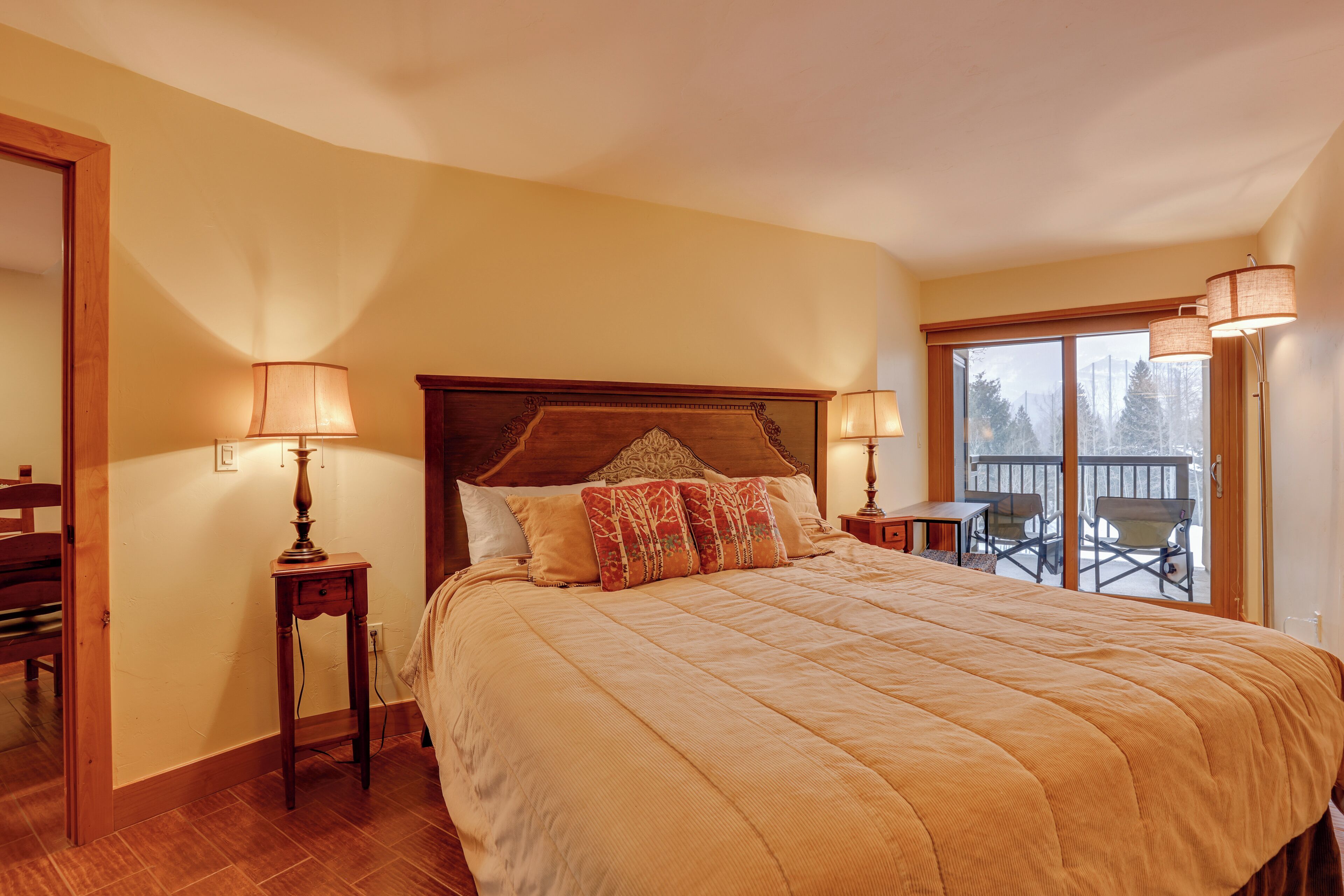 A bedroom with a large bed, two bedside lamps, a wooden headboard, and a balcony with chairs.