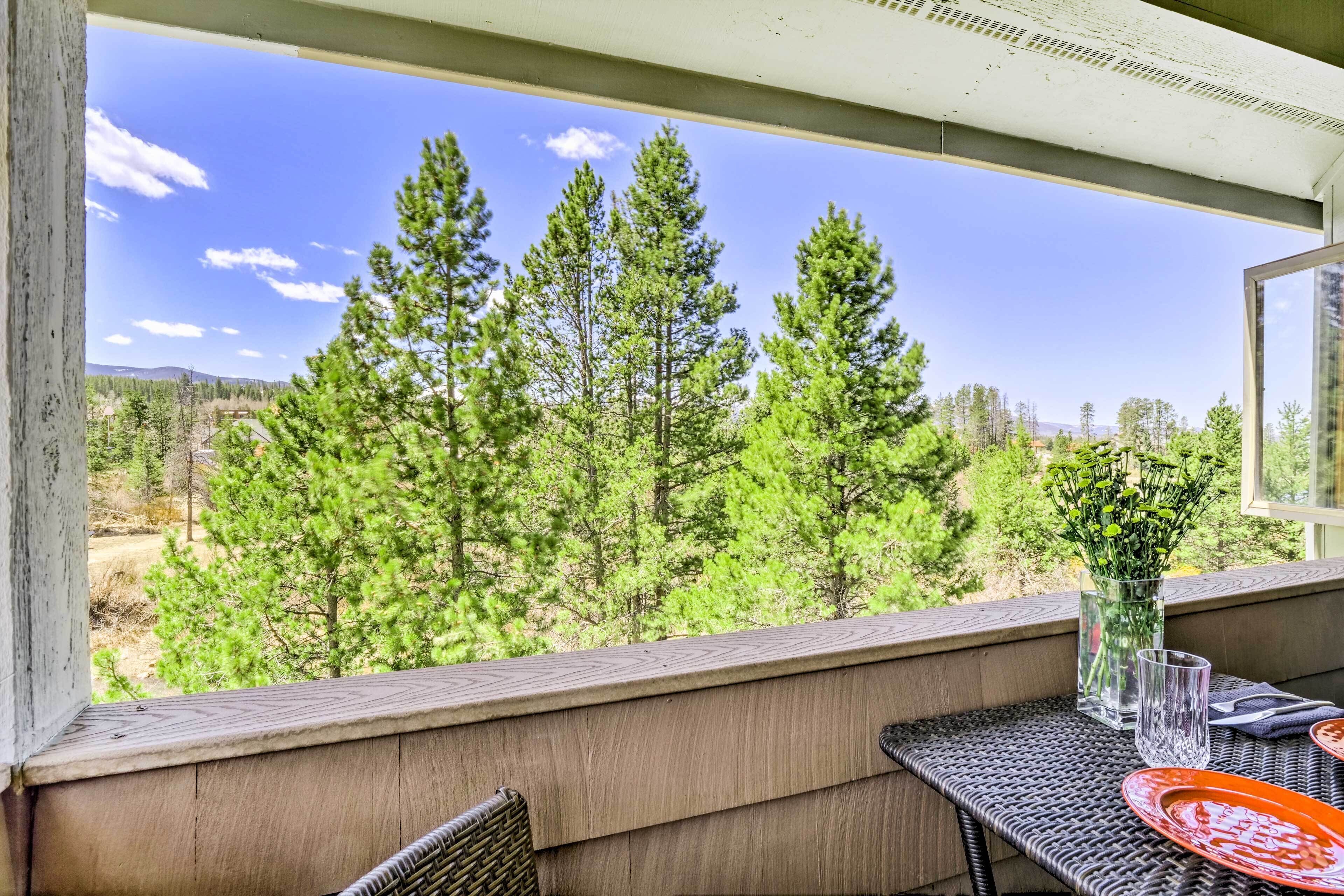 A balcony with a view of tall pine trees, a glass vase with green plants, and a table with a glass and a red plate.