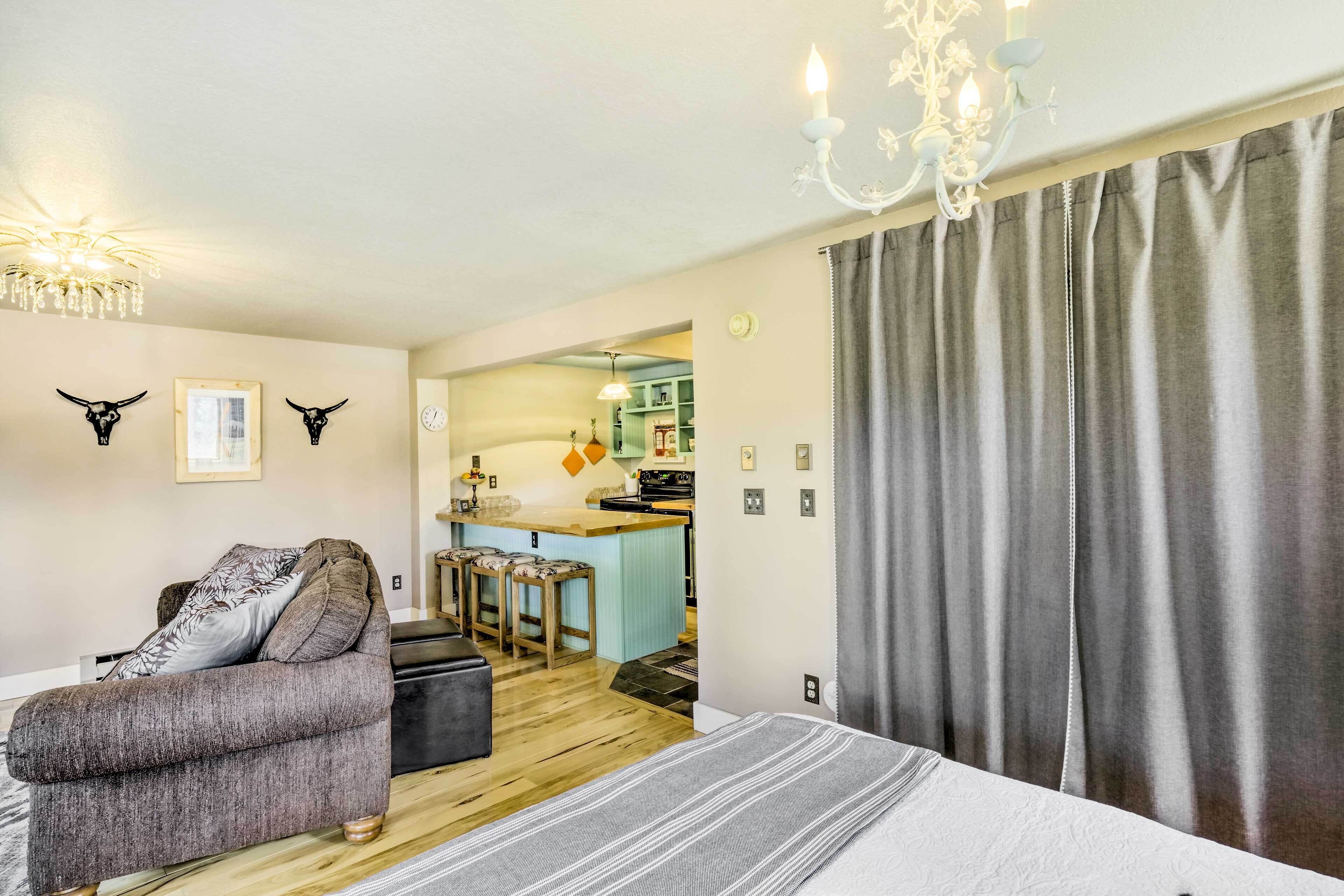 A cozy room with a sofa, a bed, a chandelier, and a kitchen area in the background.