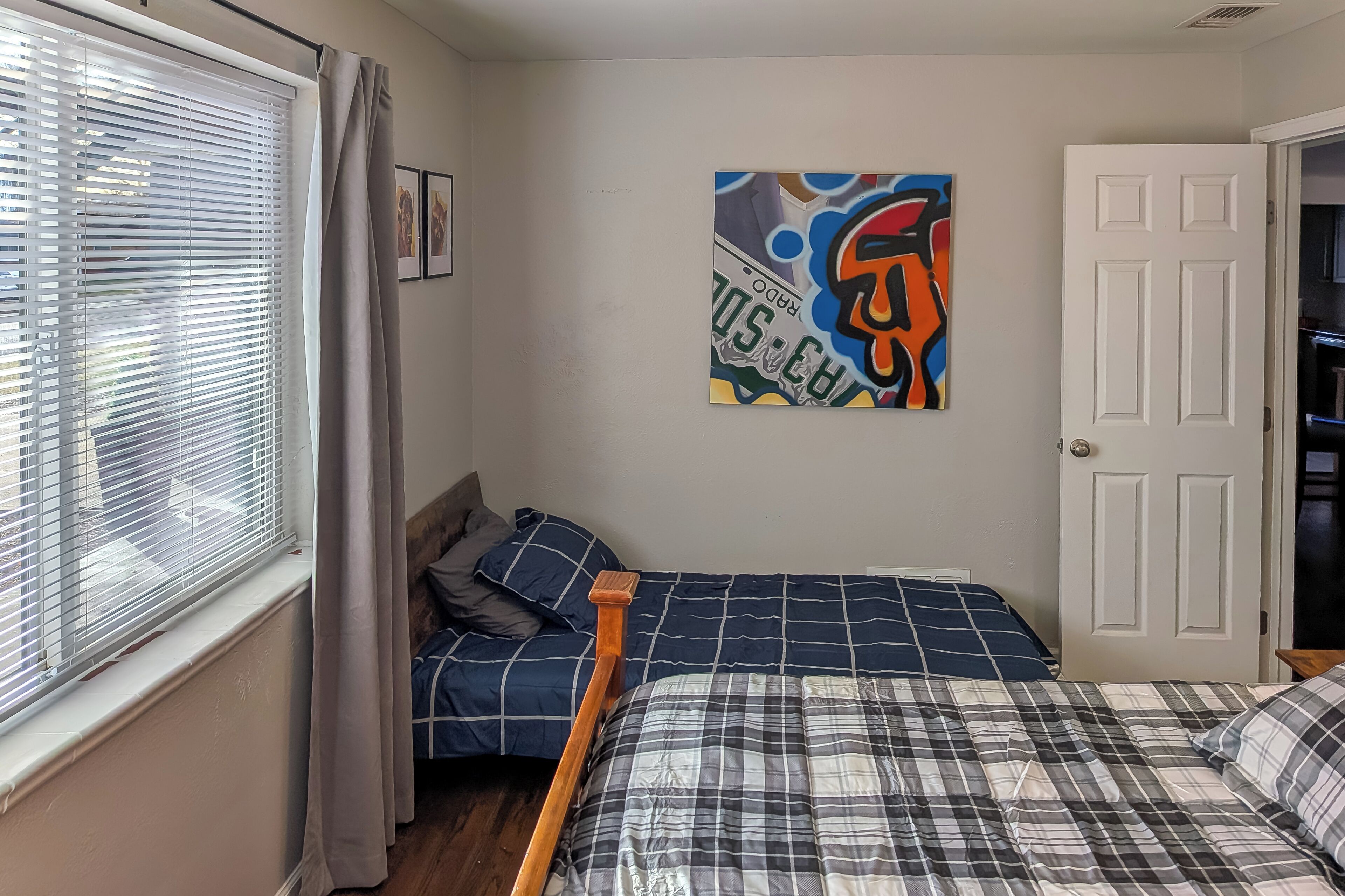 A bedroom with two beds, one with a blue and white checkered bedspread and the other with a plaid pattern. There is a window with blinds and a curtain, a door, and a wall-mounted artwork.