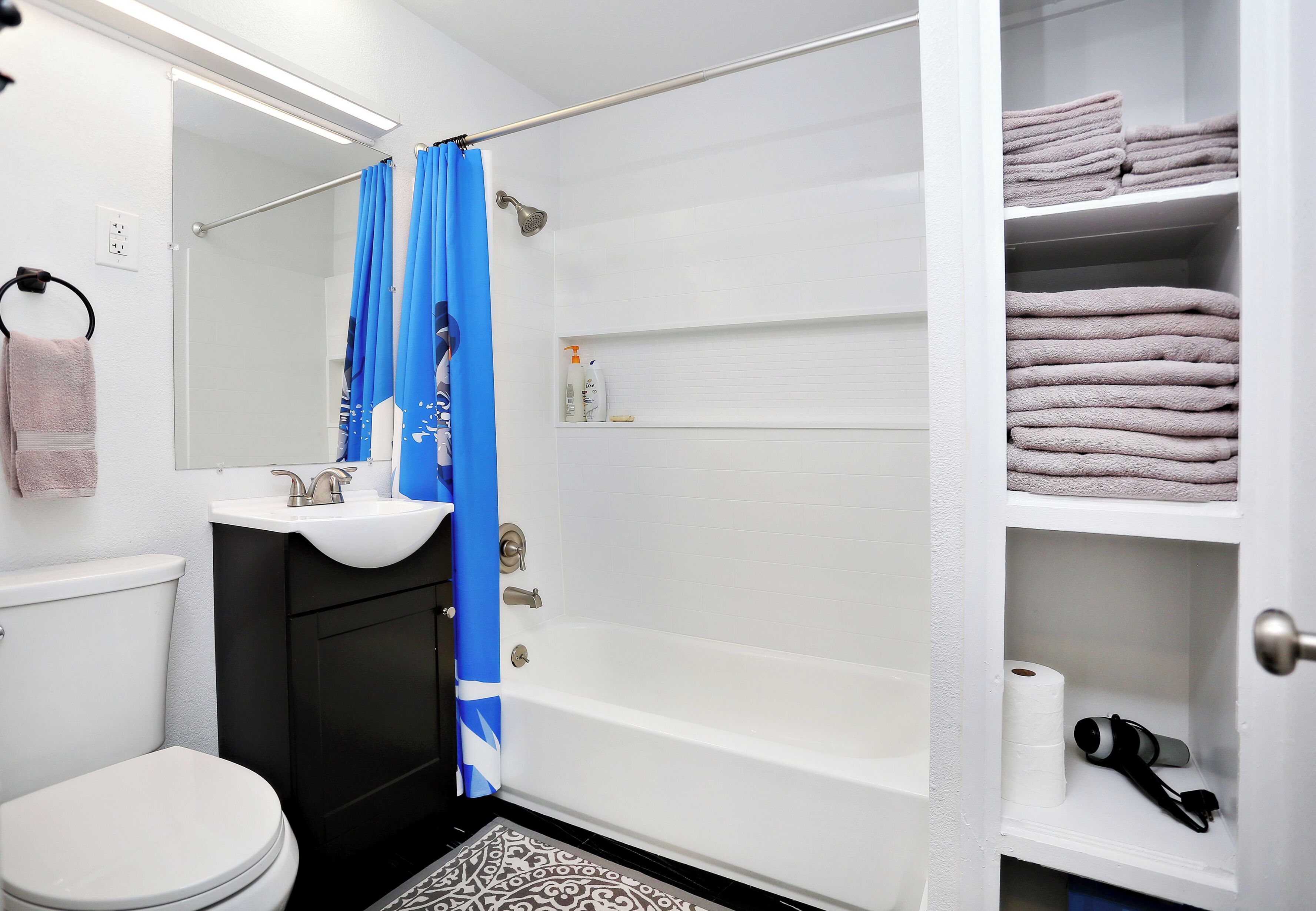 A bathroom with a white toilet, a sink, a shower with blue curtains, and shelves with towels and toiletries.