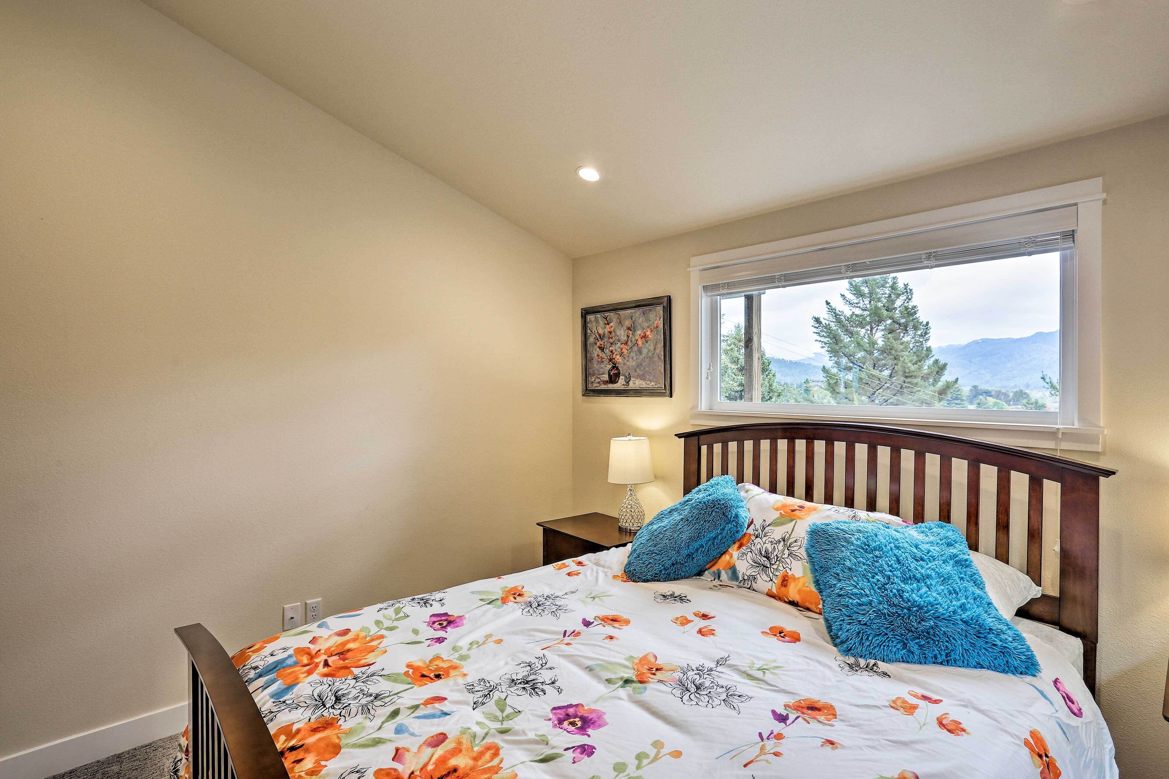 A bedroom with a floral bedspread, a wooden headboard, a nightstand with a lamp, and a window with a mountain view.