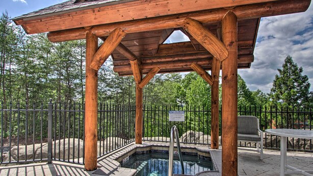 Un belvédère en bois avec un jacuzzi, entouré d’une clôture en métal et d’arbres.